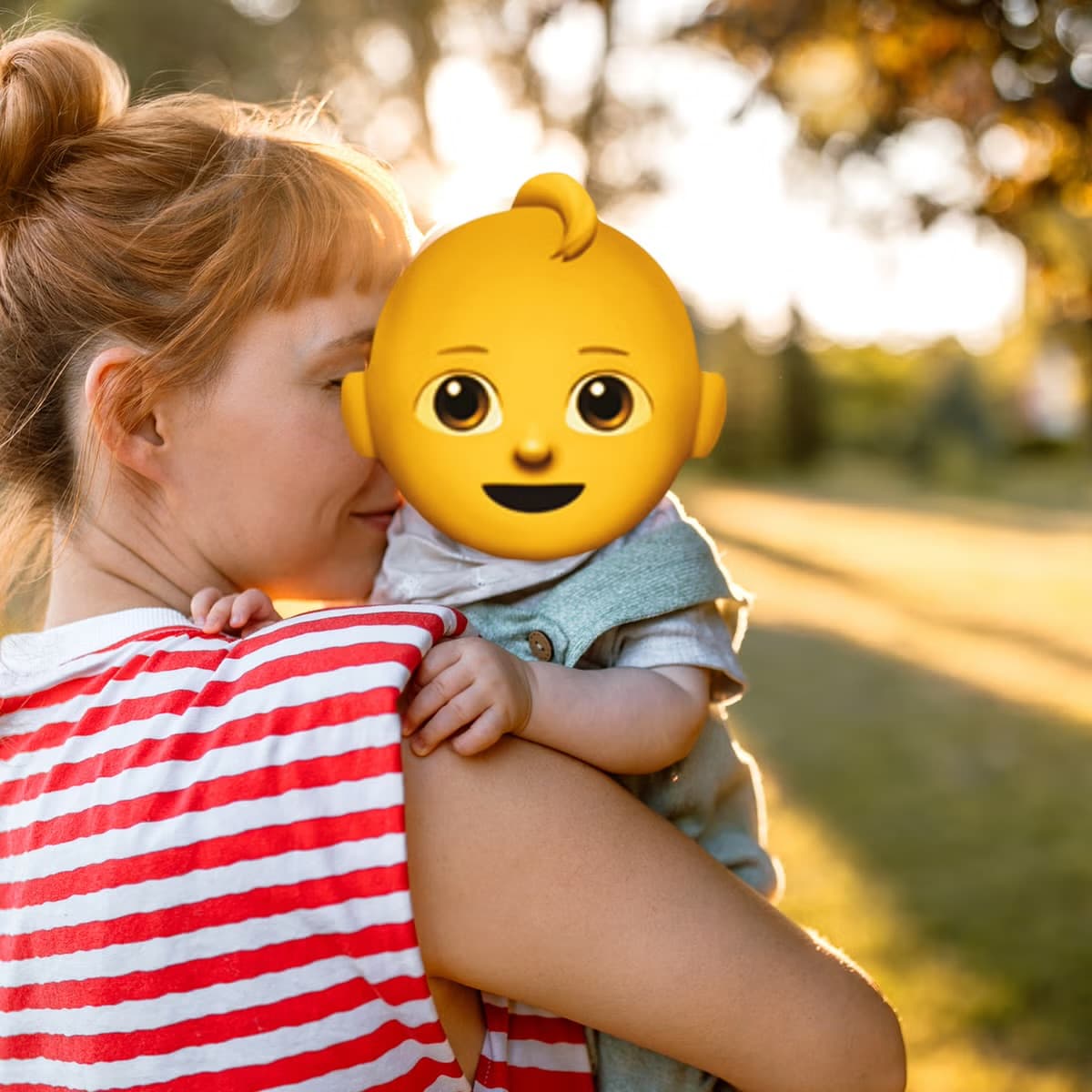 A mother holding her baby outdoors, the baby's face covered by an emoji sticker